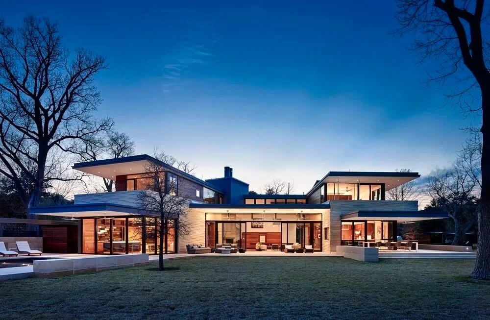 Sprawling modern home with broad overhangs glowing at dusk above a lawn and bare trees
