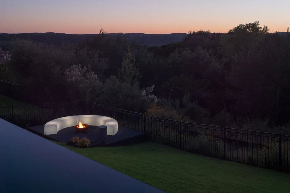 Curved glowing fire pit sofa on a lawn above the Hill Country at dusk, beside an infinity pool