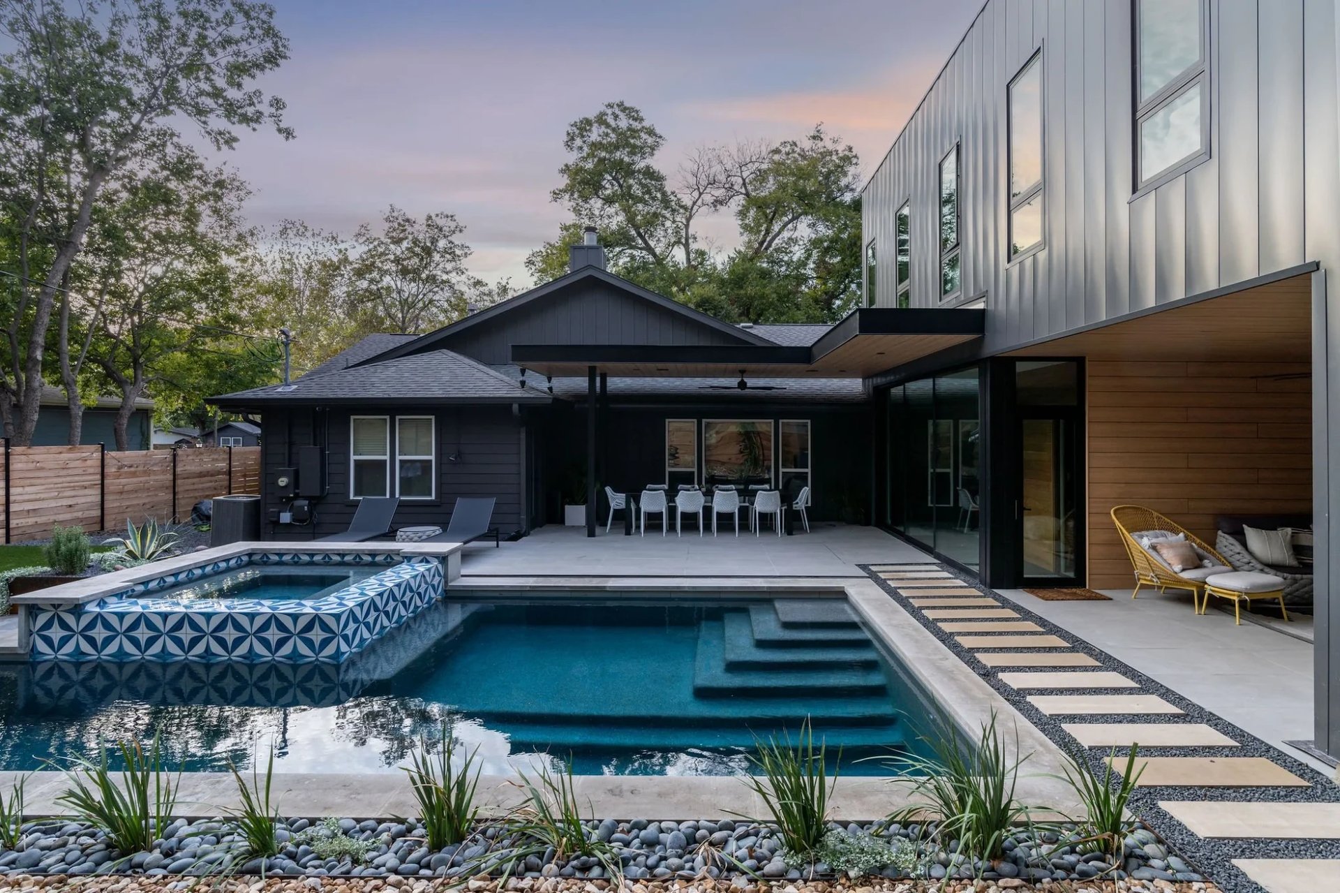 Geometric pool with blue-tile spa and steps beside a black modern home and patio at dusk in Austin