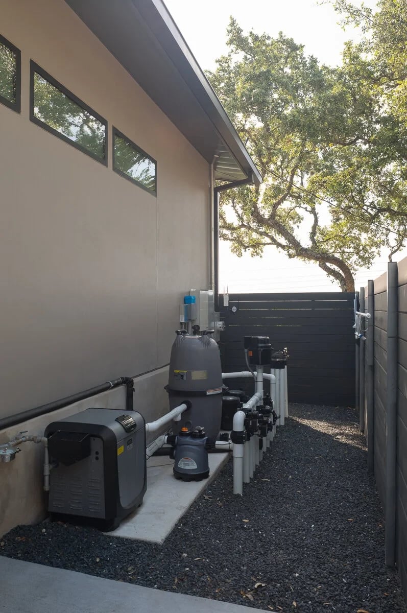 Pool equipment pad with filter and pump beside a stucco wall and gravel strip along a side yard fence