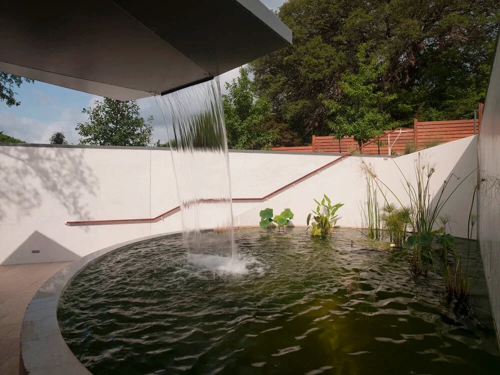 Round dark-water pool with a central fountain jet in a walled courtyard with sheer-descent spillways