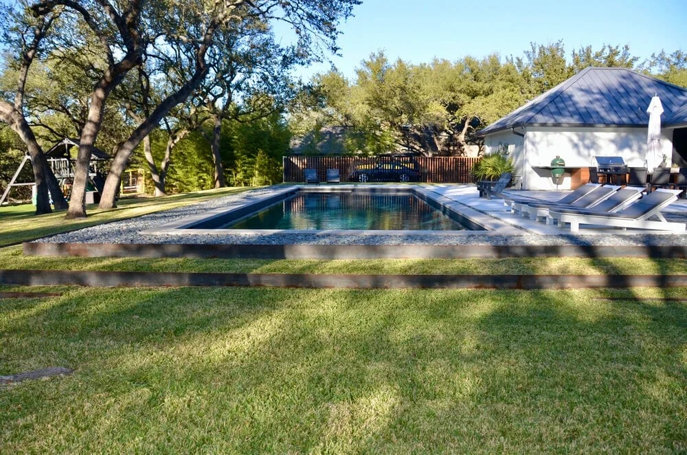Geometric pool on a lawn beyond a low rail fence, with woods and a white home behind