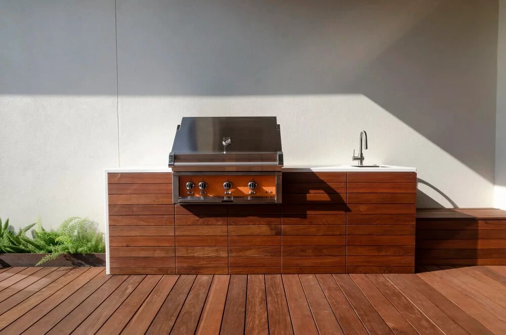 Wood-clad outdoor kitchen with a stainless grill and sink against a gray wall on an ipe deck