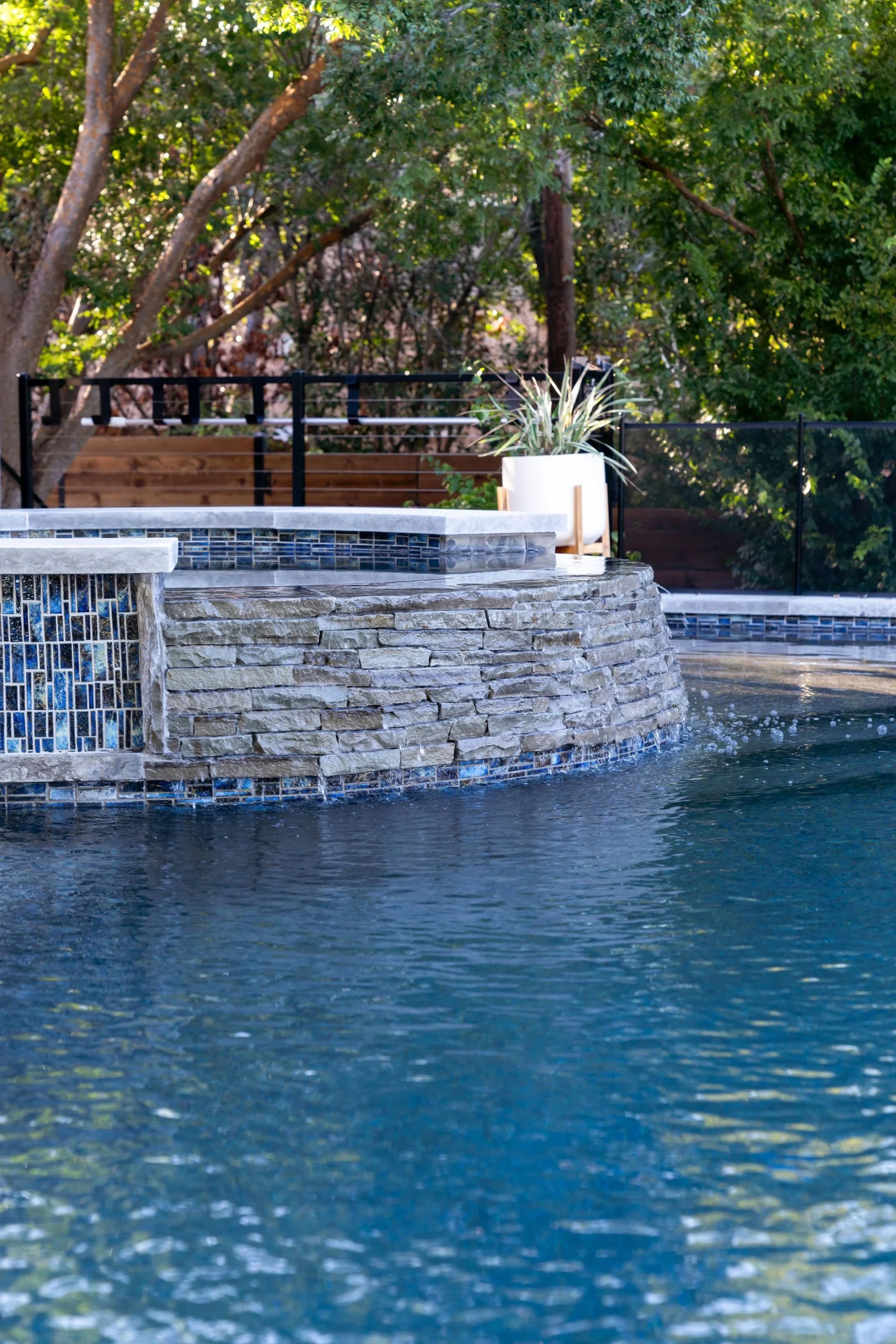 Stacked-stone spa wall with blue glass tile spilling into a pool, a potted plant and iron rail behind