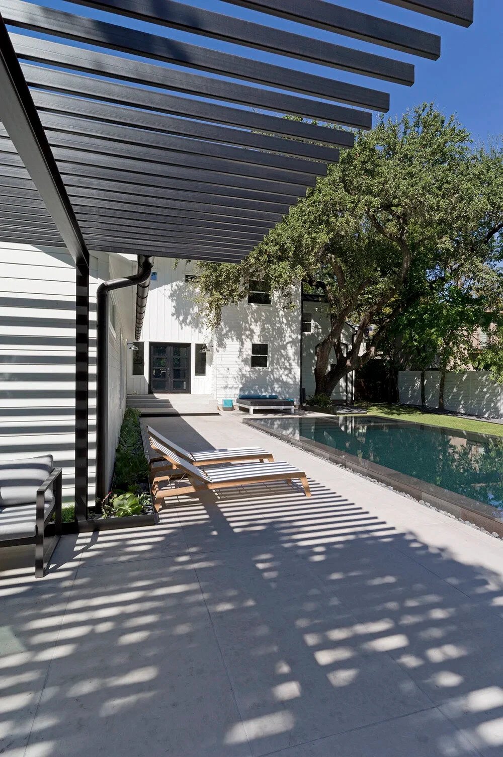 Slatted steel pergola shading teak loungers along a long dark lap pool beside a white modern home