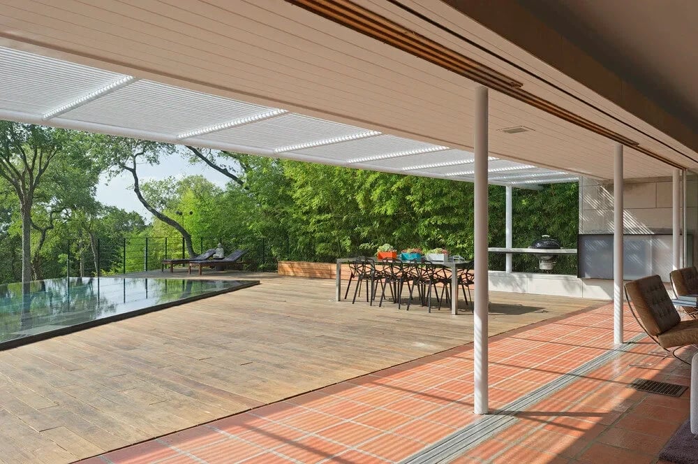 Covered patio with a slatted ceiling, dining table and wood deck beside an infinity pool among trees