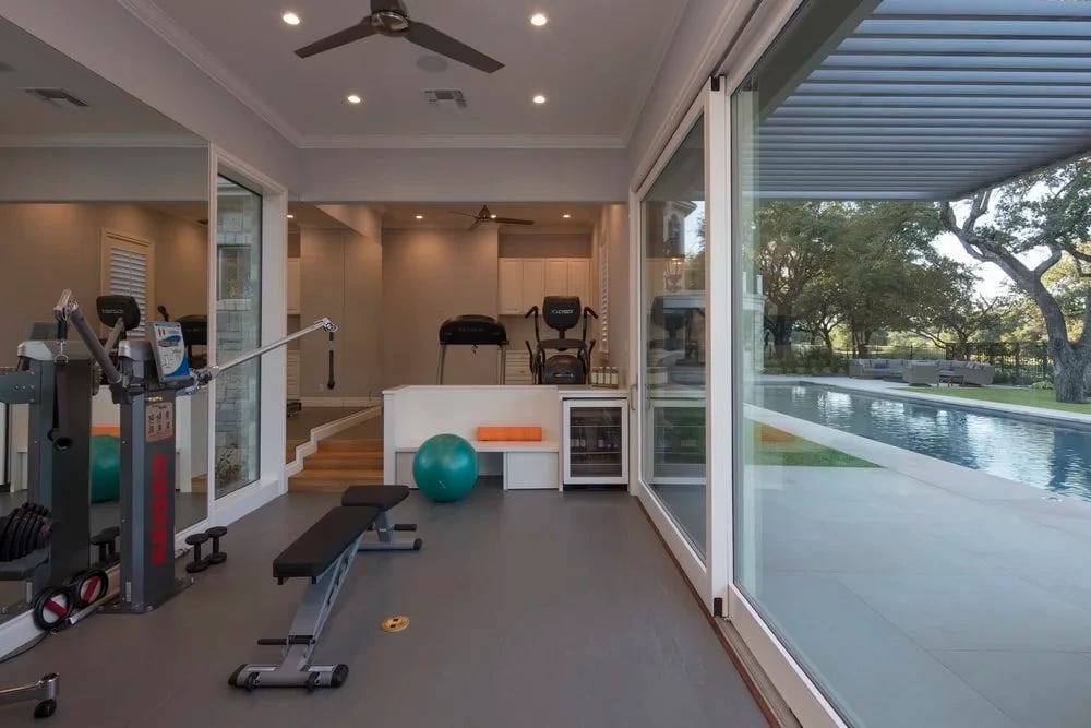Indoor home gym with sliding glass walls opening onto a geometric pool and oak-shaded lawn