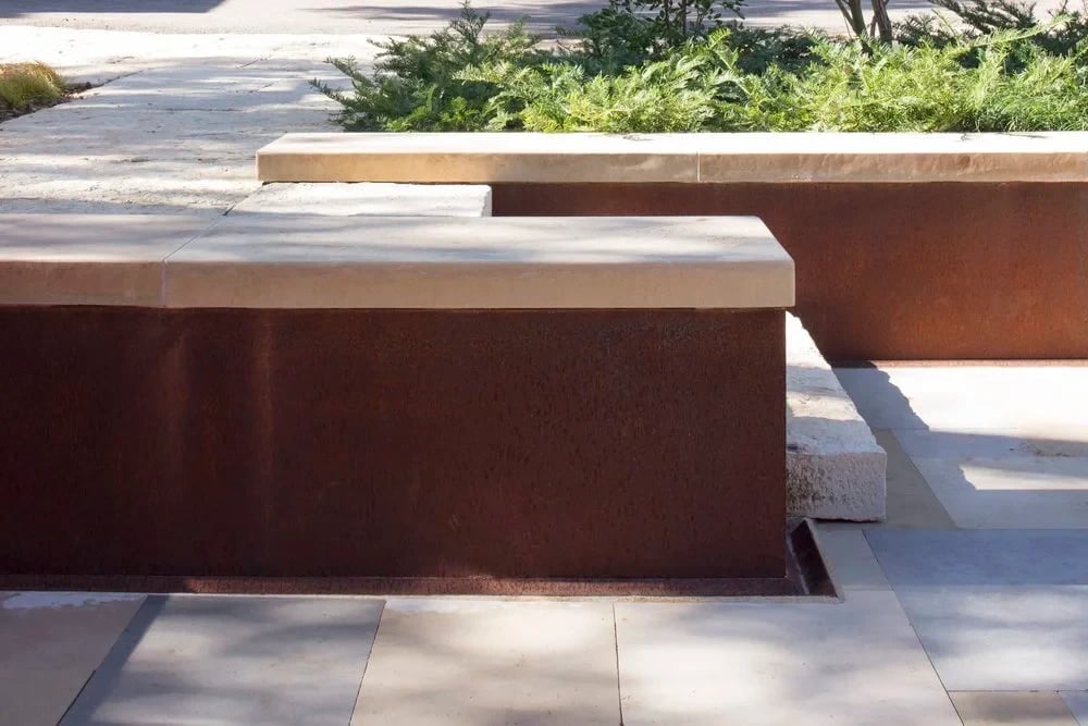 Weathered steel planter benches with stone caps along a paved walkway beside green plantings