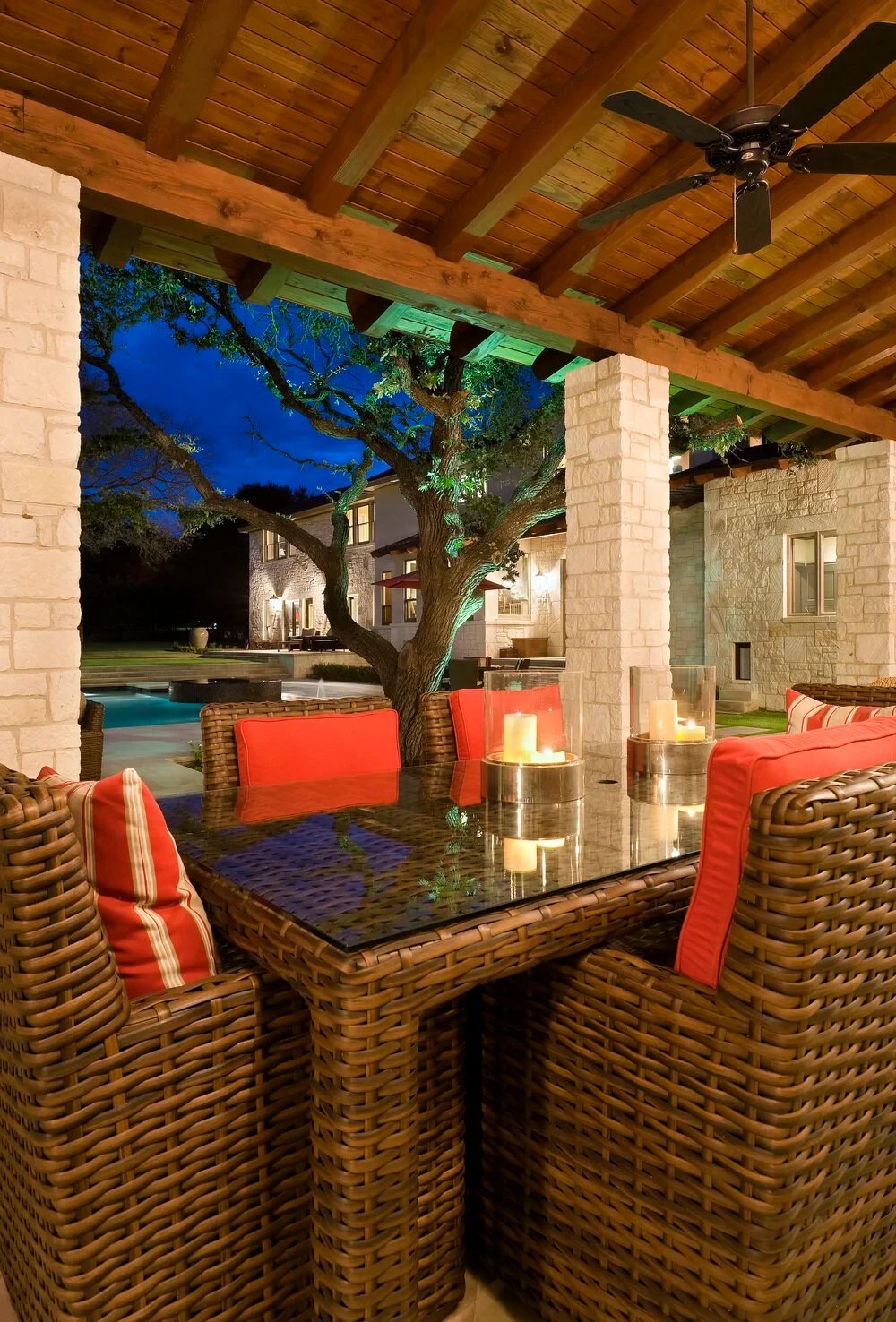 Covered patio at dusk with woven chairs, a candle table, and a lit pool and stone home beyond