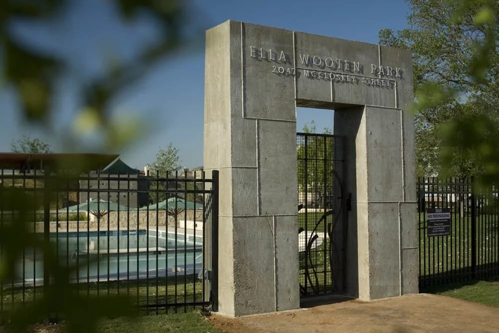 Concrete gateway reading Ella Wooten Park beside a fenced swimming pool, viewed through green leaves
