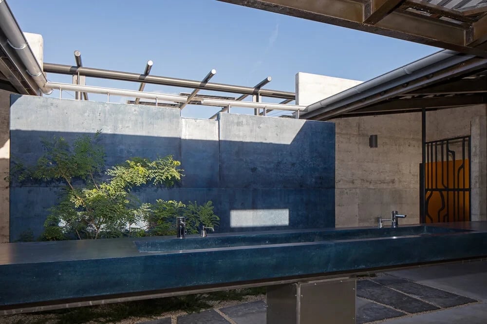 Concrete bathhouse courtyard with steel pergolas and a blue panel beside a still pool