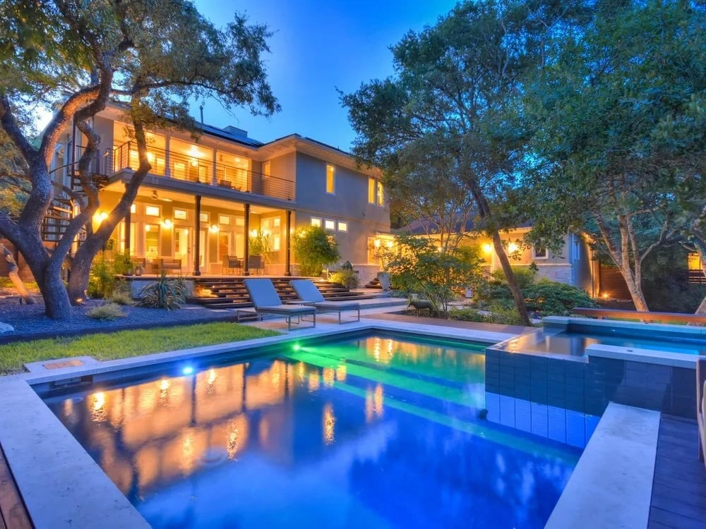 Geometric pool and spa lit blue and green at dusk beside a glowing two-story home under live oaks