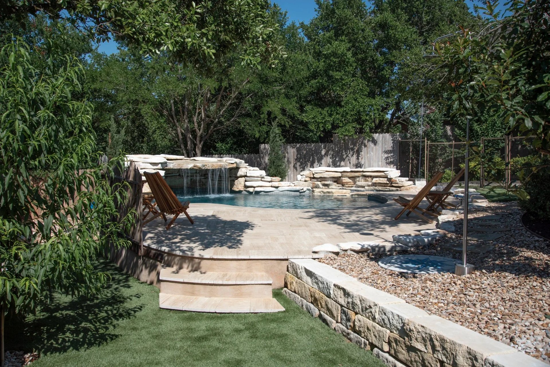 Freeform pool with stone waterfall, teak chairs and a pebble-filled spa beside a travertine deck