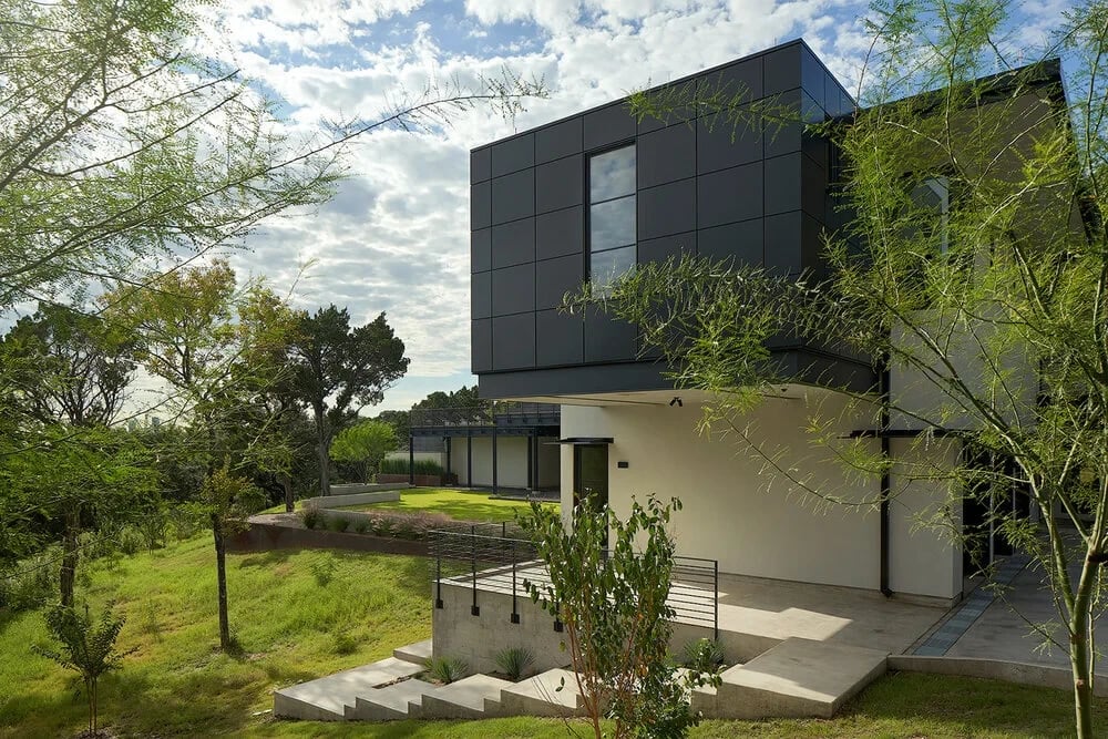 Dark-clad modern home cantilevered over a hillside, framed by trees and native grasses