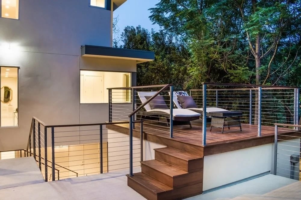 Raised wood deck with lounge chairs and cable railing beside a modern stucco home at dusk