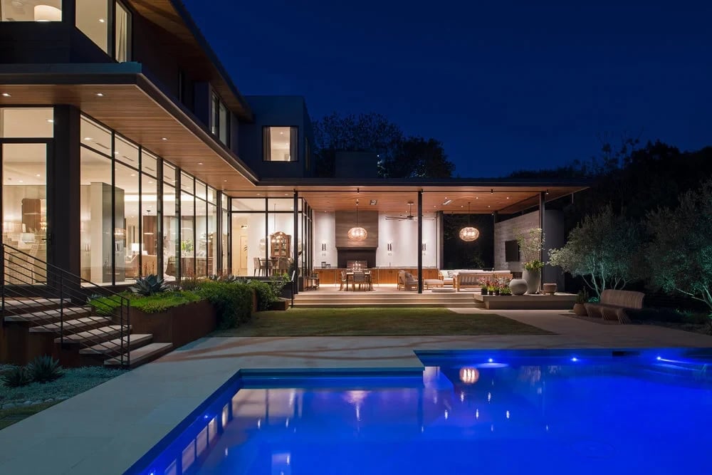 Modern glass-walled home with a lit outdoor kitchen glowing at dusk above a geometric pool with blue lighting