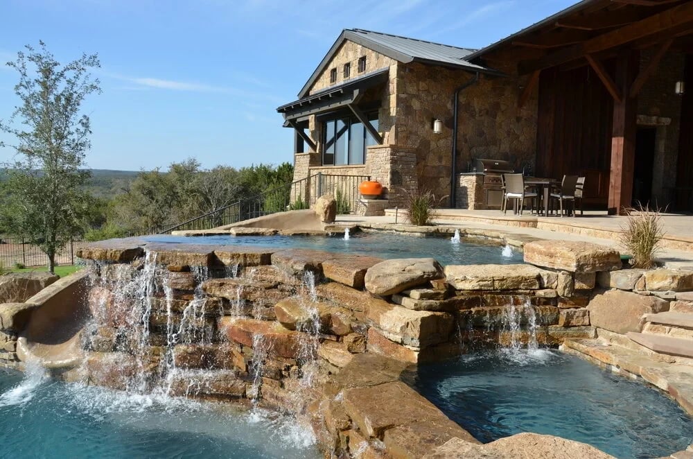 Freeform pool spilling over layered stone waterfalls below a stone home with a covered patio and Hill Country view