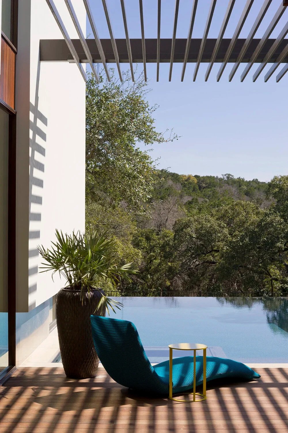 Infinity pool edge vanishing over a wooded hillside, with a lounger and potted palm on a wood deck