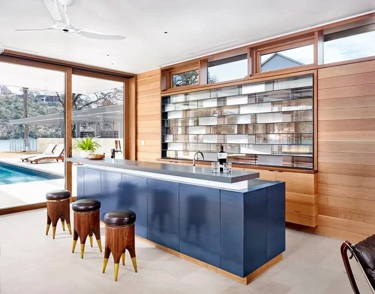 Wood-paneled kitchen with a navy island and wood stools, glass wall opening to the pool