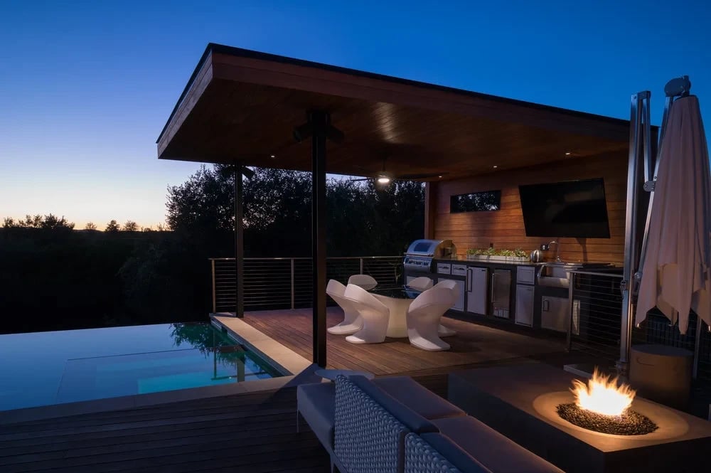Lit cabana with outdoor kitchen and white chairs beside an infinity pool and fire pit at twilight