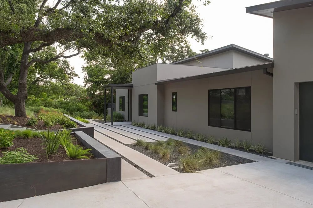 Modern stucco home entry with a low-water garden, board-formed planters and a paved walkway under live oaks