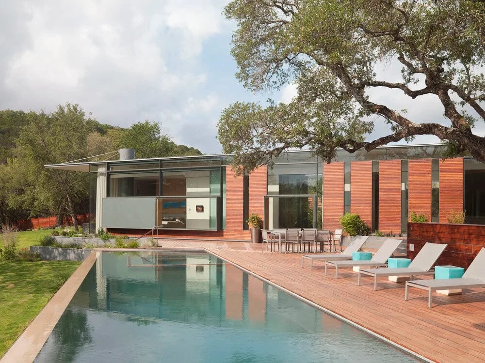 Geometric pool beside a wood deck with lounge chairs and a glass-walled home under live oaks