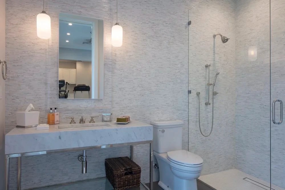 Marble-topped vanity and glass walk-in shower in a bath tiled with pale linear stone
