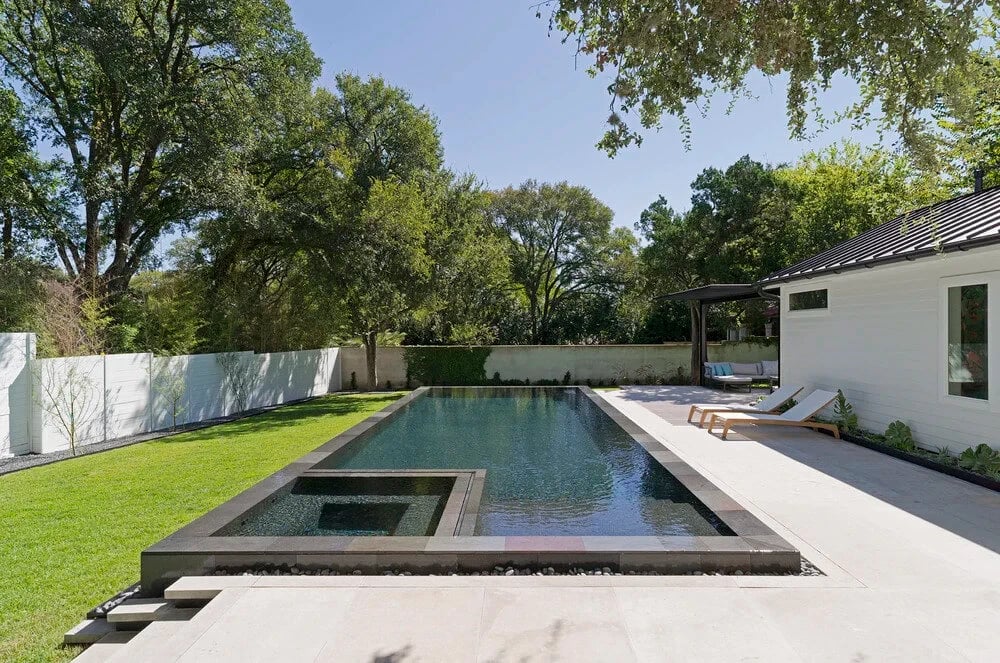 Dark geometric pool with a perimeter-overflow edge and a spillway spa in a fenced, tree-lined backyard