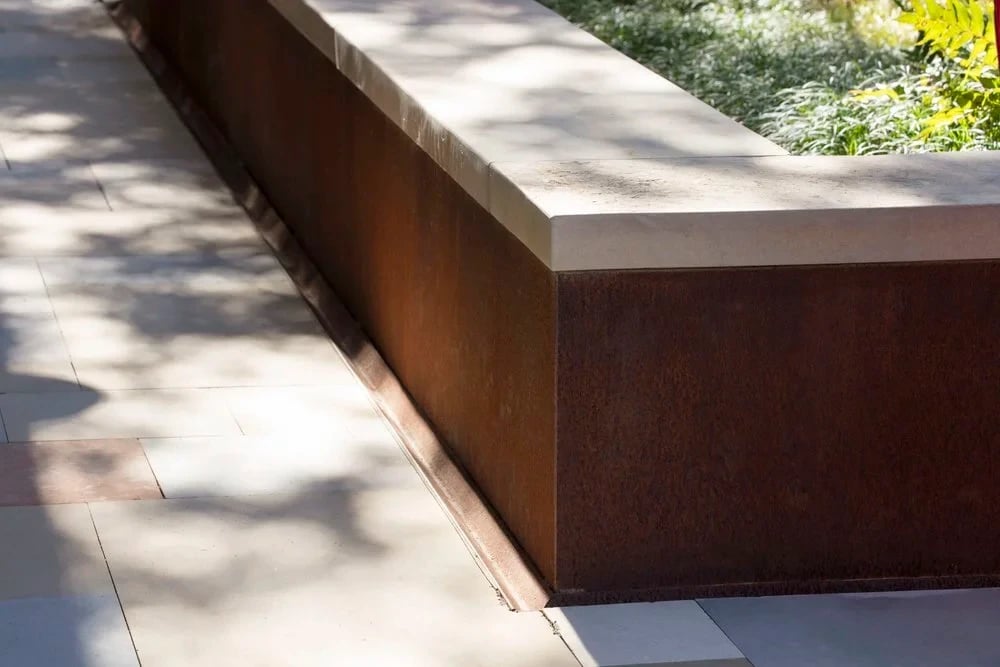 Stone-capped weathered steel retaining wall casting dappled shade over a paved path