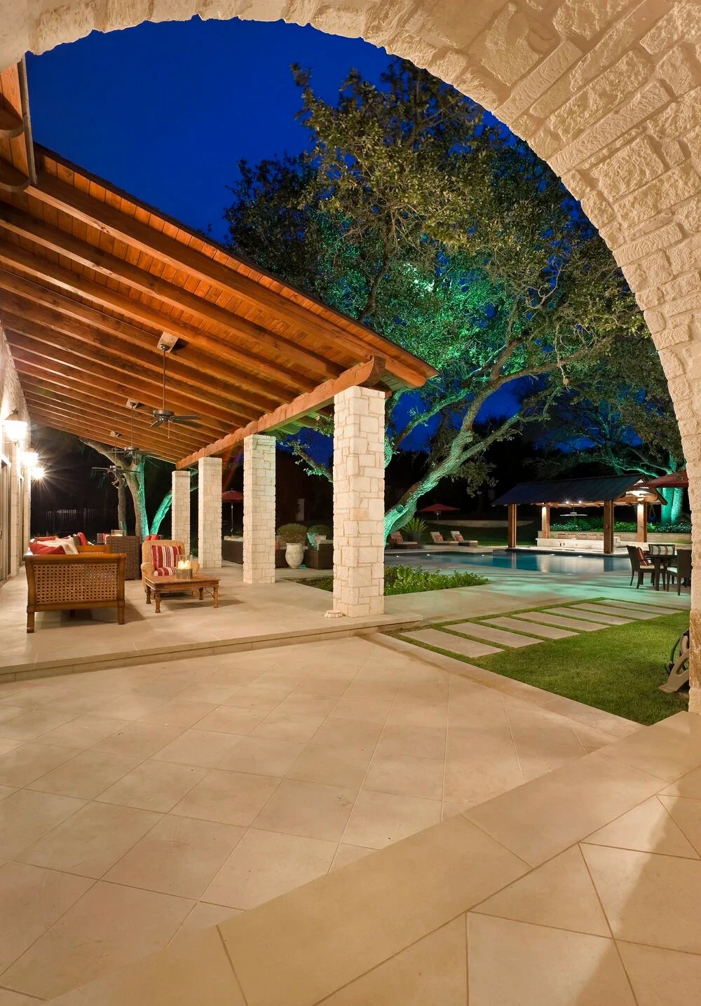 Stone arch framing a covered limestone patio with seating and a lit pool and lawn at twilight