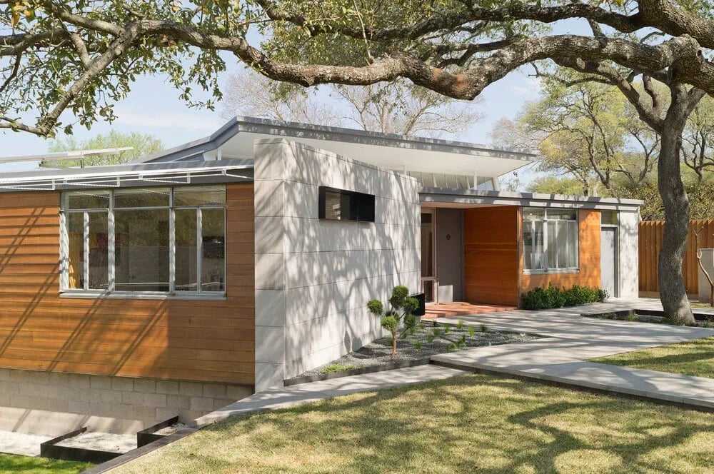 Single-story modern home clad in cedar and white stone, shaded by live oaks over a stone walkway