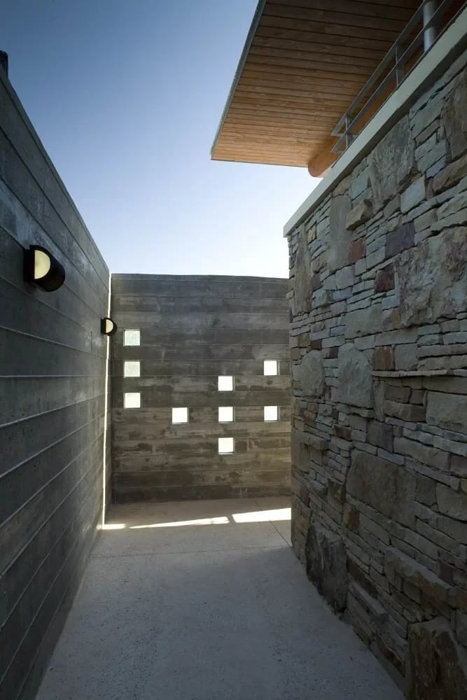 Narrow passage between board-formed concrete and stone walls, glass-block wall lit by a clear sky
