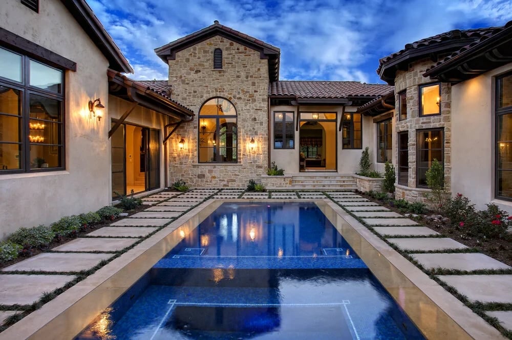 Stone Mediterranean home reflected in a geometric courtyard pool, paver-and-grass deck at twilight