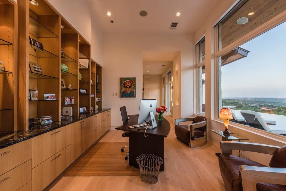 Home office with wood built-in shelves, a dark desk, and tall windows over the Hill Country at dusk