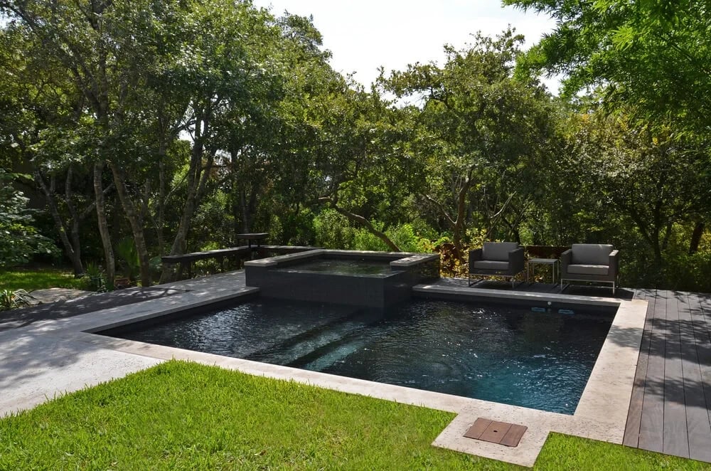 Dark geometric pool with a raised spa, stone coping, and a green lawn ringed by leafy trees