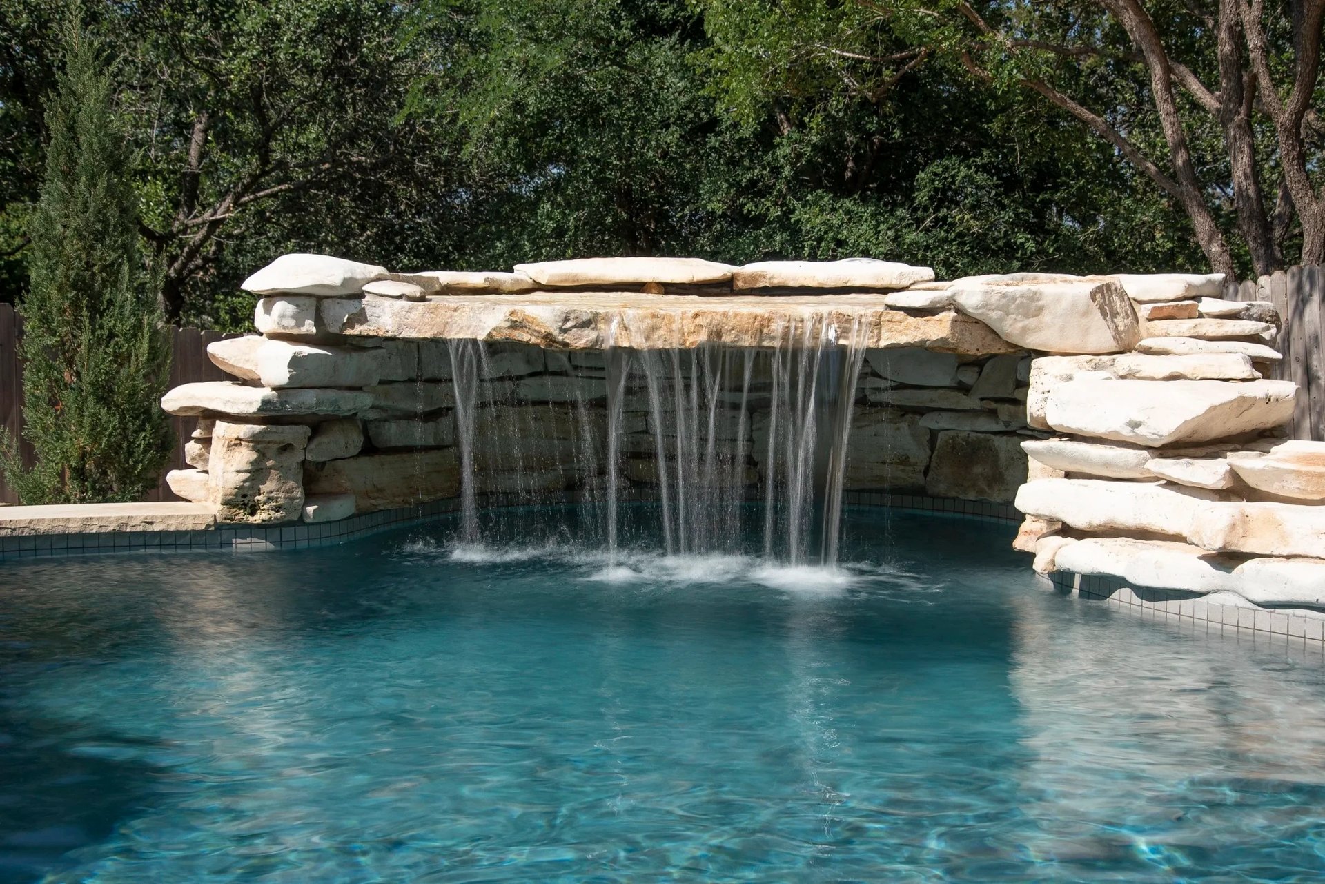 Stacked-stone waterfall spilling into a turquoise freeform pool framed by cedars and a privacy fence