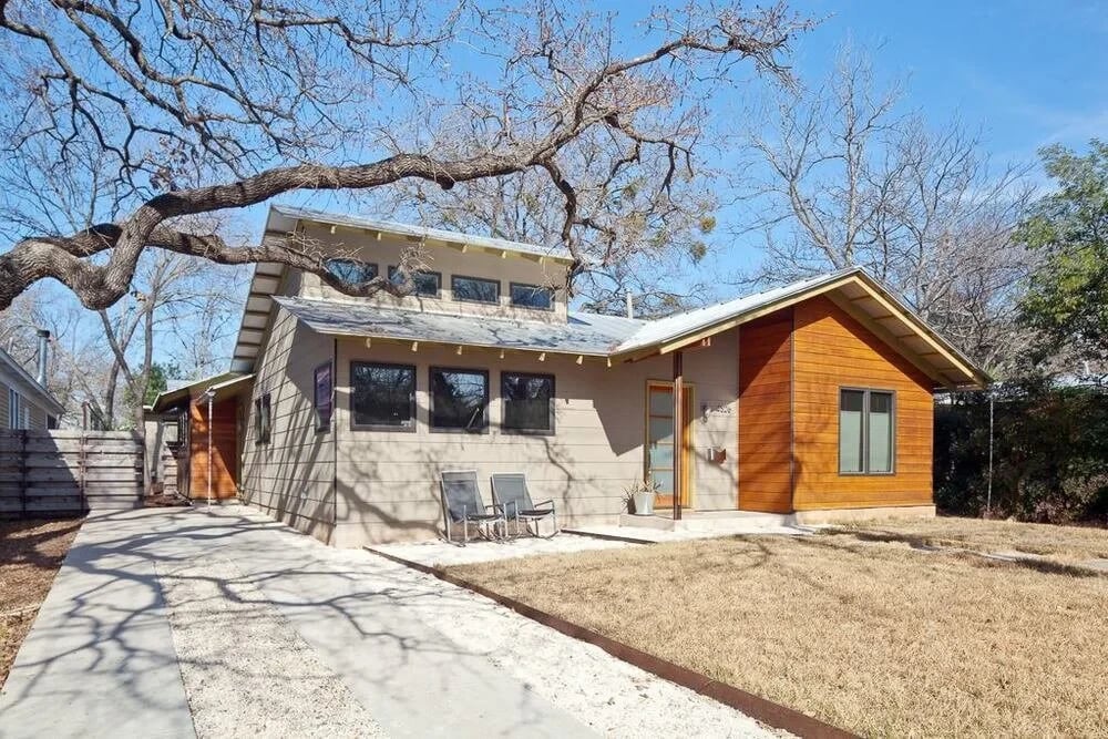 Modern stone-and-cedar home with clerestory windows behind a bare oak on a gravel drive