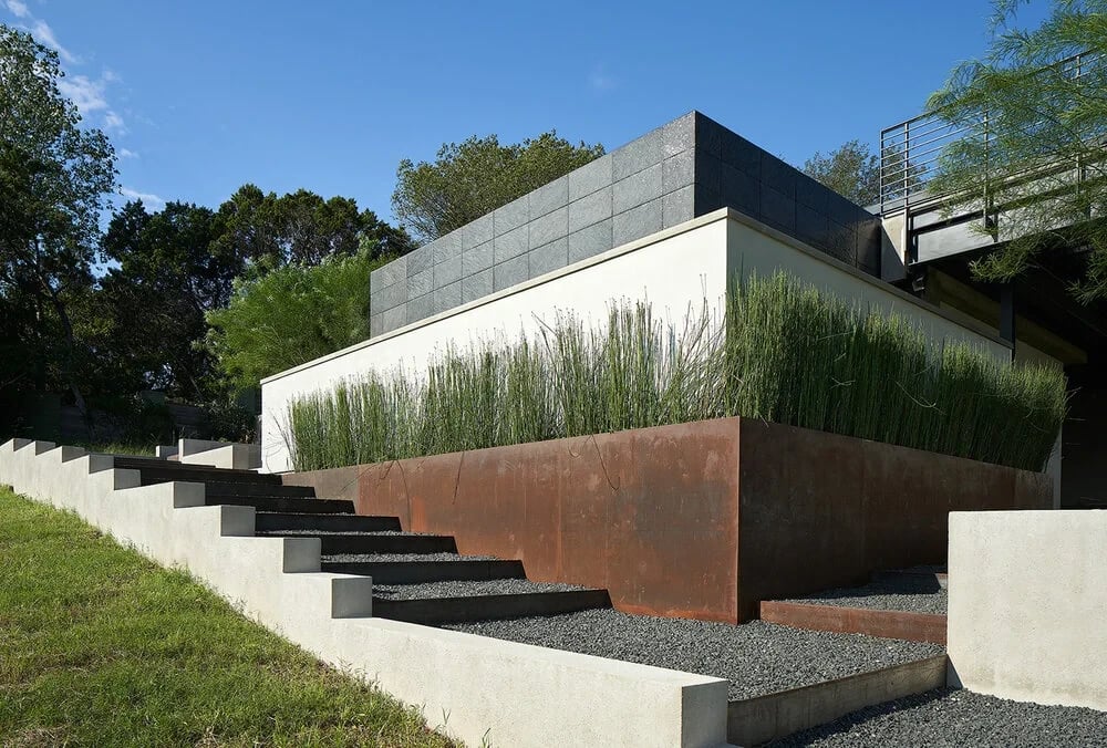 Modern home with weathered steel walls and grasses above concrete steps set in gravel