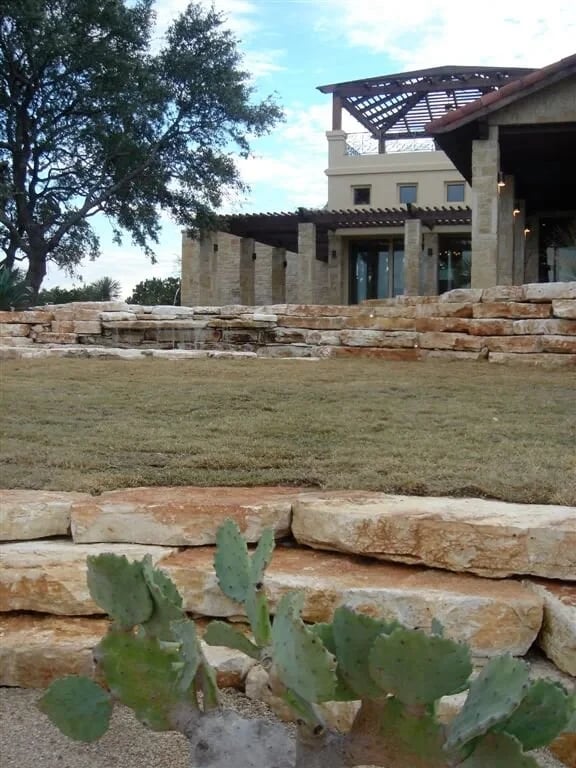 Stone lodge with a pergola above tiered limestone walls, lawn and prickly pear in the foreground