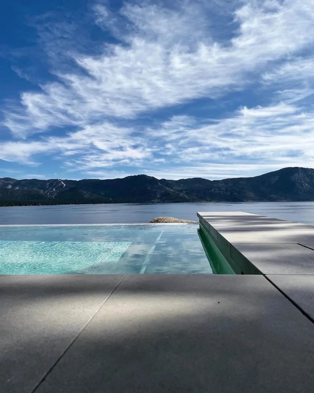 Infinity pool edge meeting a calm lake and forested mountains under a wide blue sky with wispy clouds