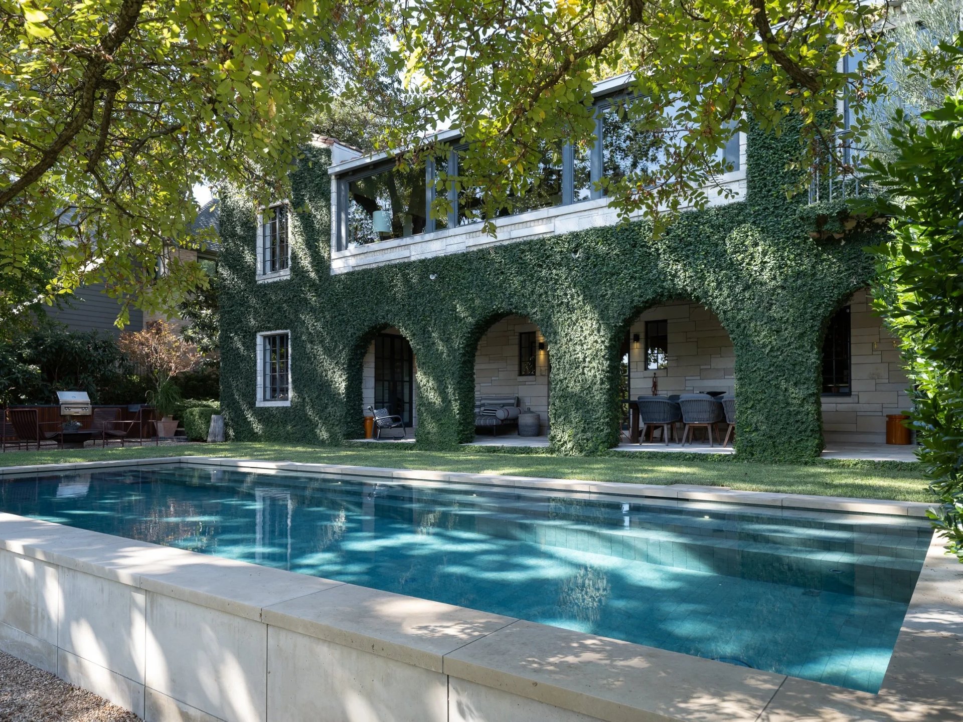 Geometric pool beside a two-story ivy-covered home with arched openings and a shaded lawn