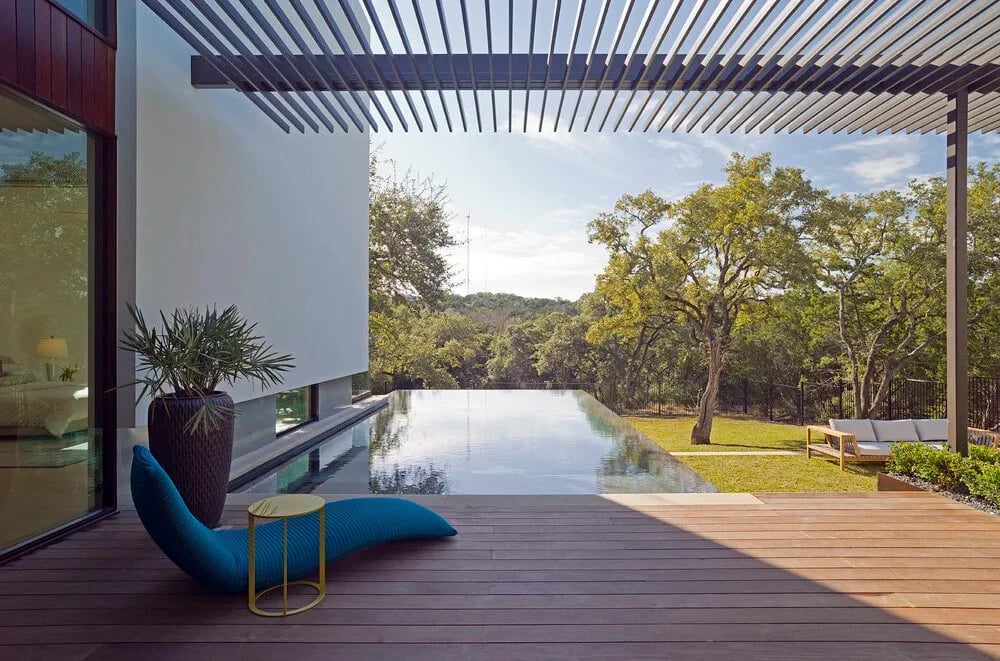 Slat pergola over a wood deck and infinity pool overlooking trees, with a blue lounger and palm