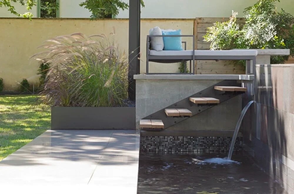 Detail of a poured-concrete spa with floating wood steps, mosaic tile and a scupper spilling into water