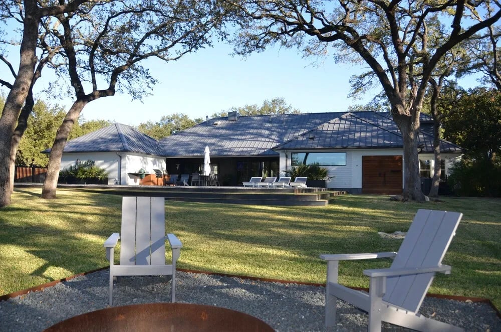 Adirondack chairs and a fire pit on a lawn facing a white metal-roof home under oaks