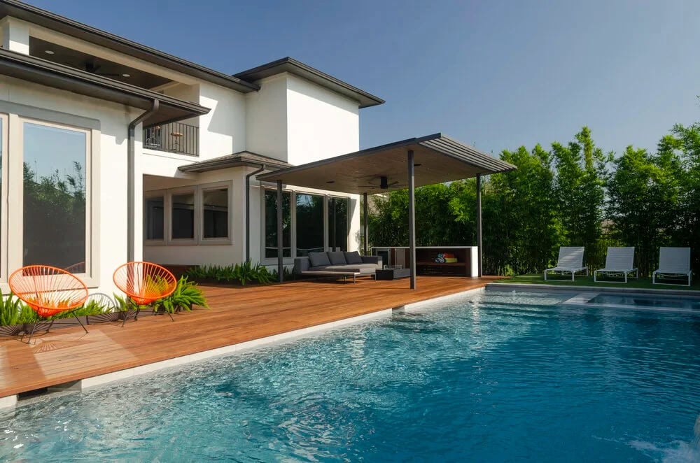 Geometric pool with an ipe deck and covered patio beside a two-story modern home and bamboo hedge