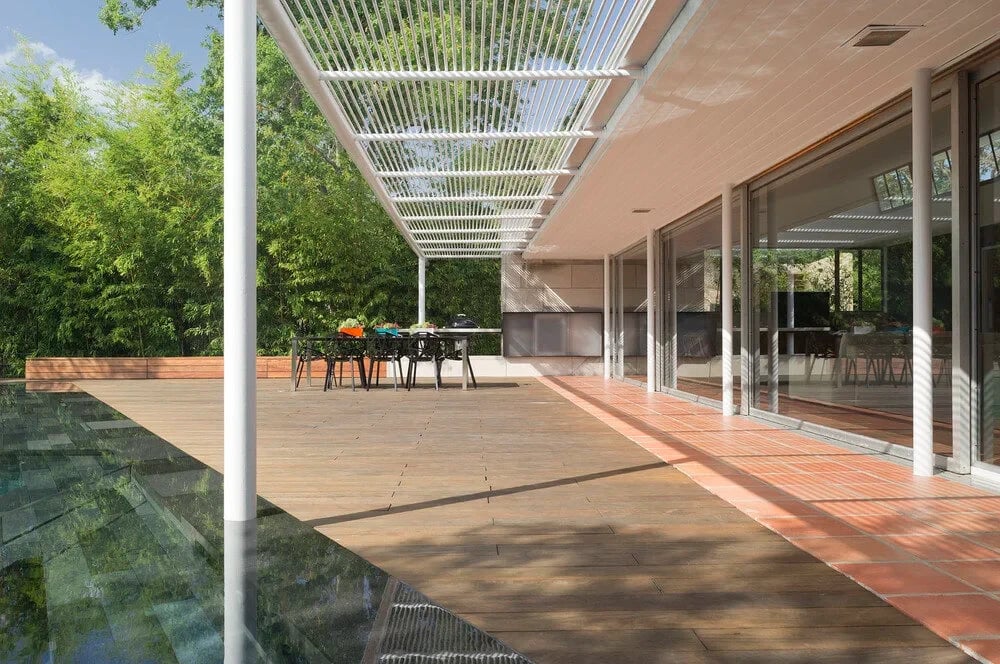 Wood-deck patio under a white pergola with a dining set, infinity pool and woods alongside
