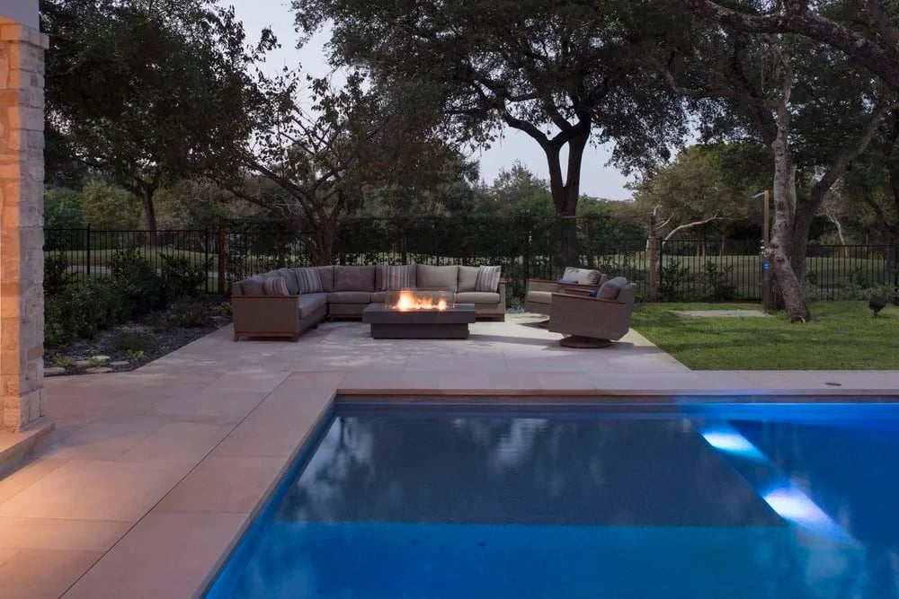 Patio sectional and gas fire table beside a geometric pool, fenced lawn and oaks beyond at dusk