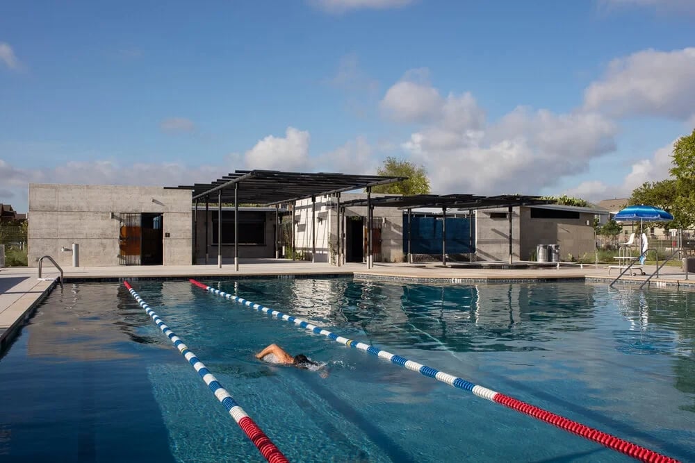 Swimmer doing laps in a lane-lined public pool before a concrete bathhouse and steel pergolas
