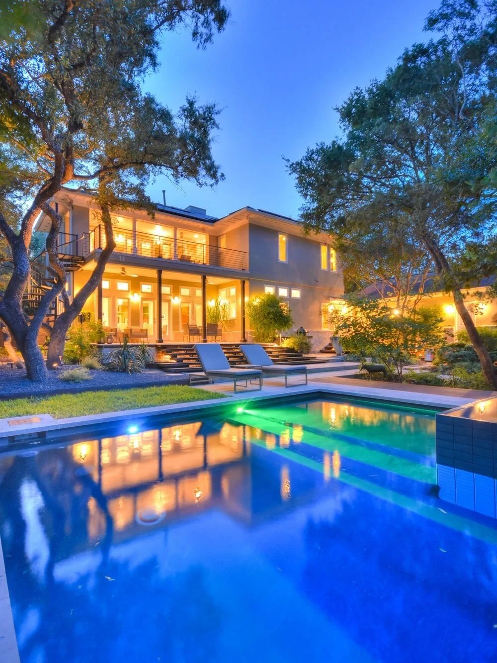 Geometric pool lit blue and green at twilight below a glowing two-story home framed by oak trees