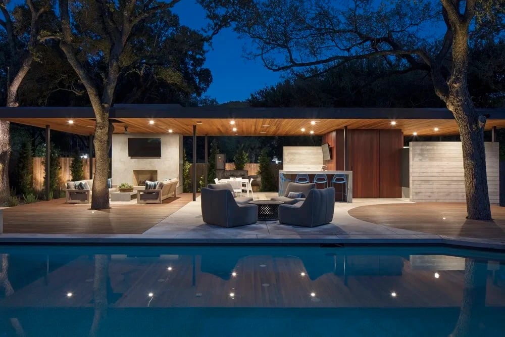 Lit pool at dusk reflecting a covered patio with a fireplace, lounge seating and an outdoor kitchen